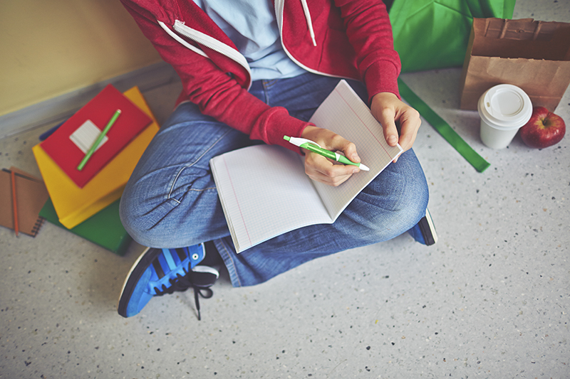 Schoolboy in casualwear making notes in exercise-book while sitting on the floor